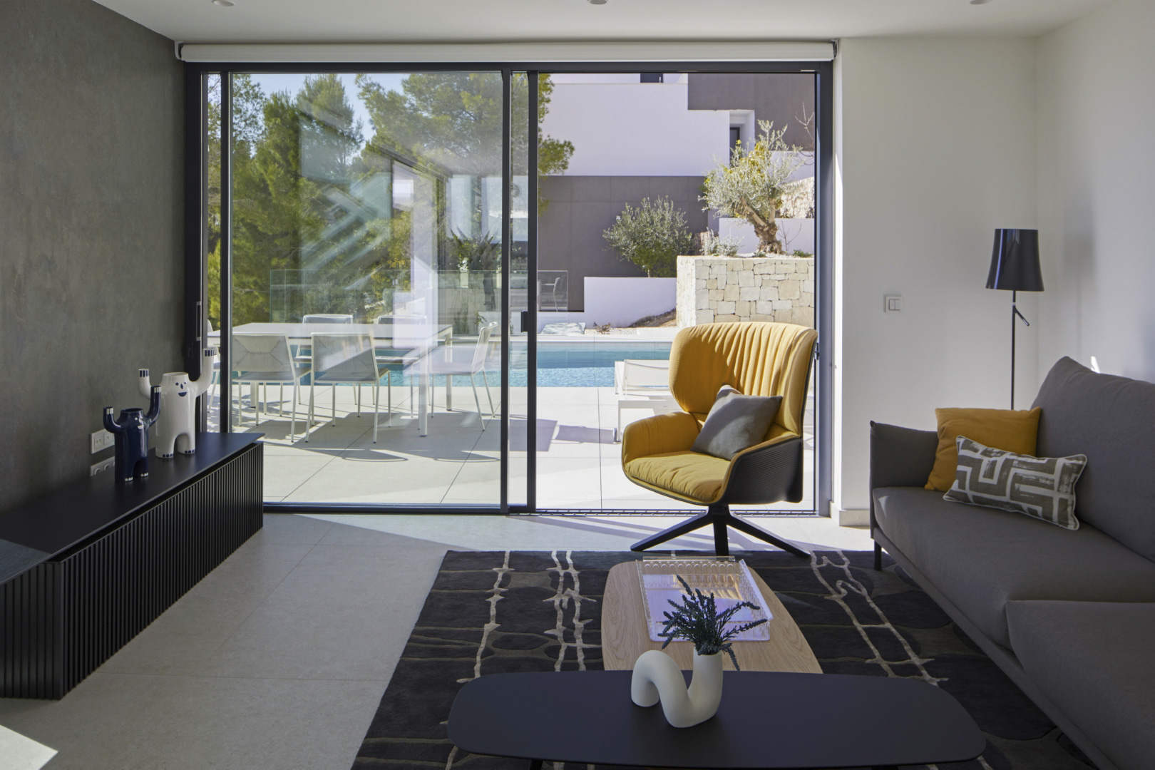 Salón moderno con amplios ventanales correderos que conectan con la terraza y la piscina. Incluye mobiliario de diseño, una butaca amarilla y suelos de gres porcelánico.