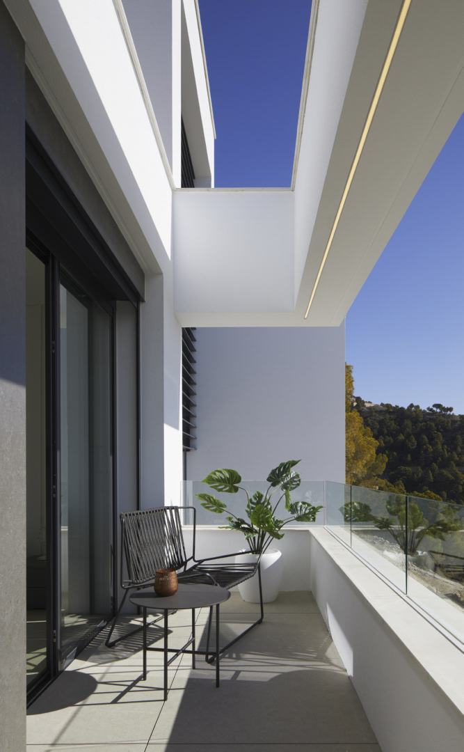 Terraza privada con barandilla de cristal, mobiliario de exterior minimalista y vistas a la montaña. Incluye iluminación técnica integrada en el voladizo blanco.