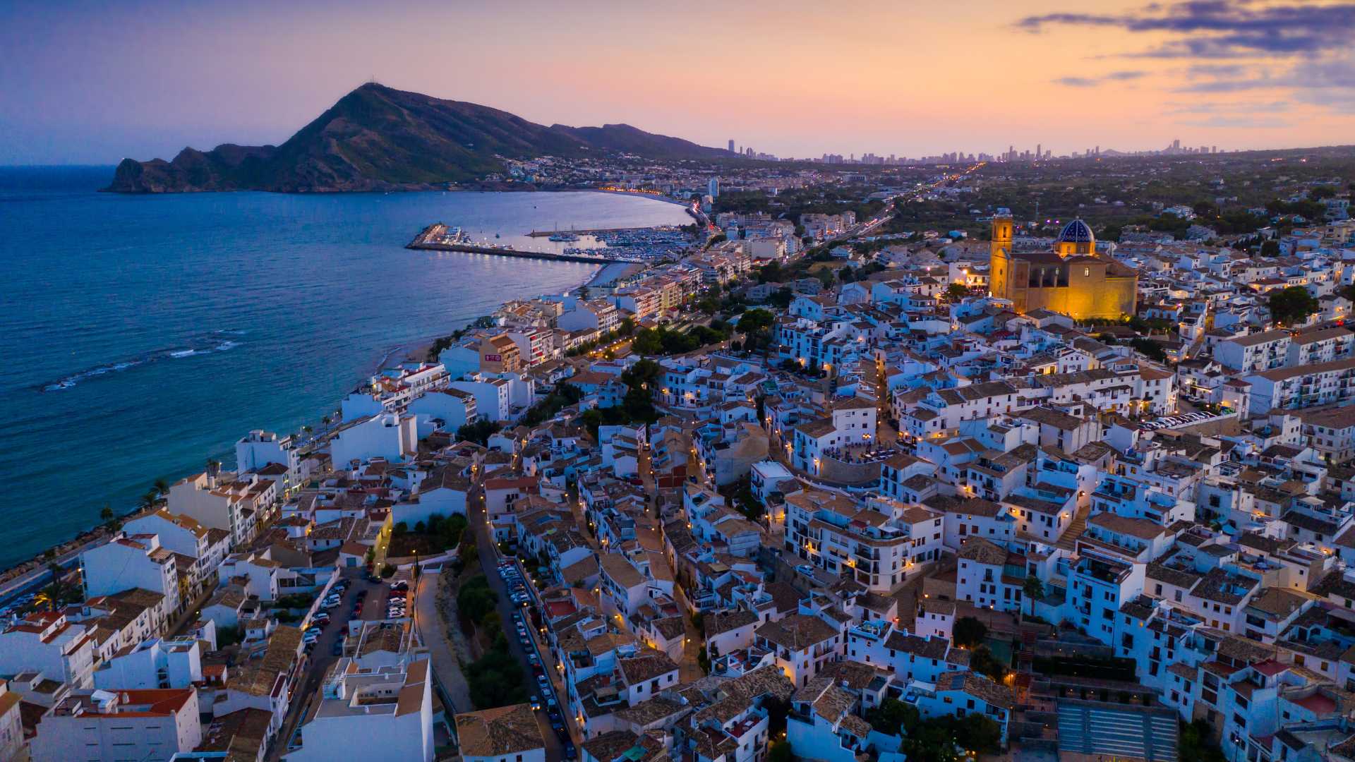 Vista aérea crepuscular del casco antiguo de Altea, destacando la iglesia de cúpula azul, la costa mediterránea y la silueta de la Sierra Helada al fondo.