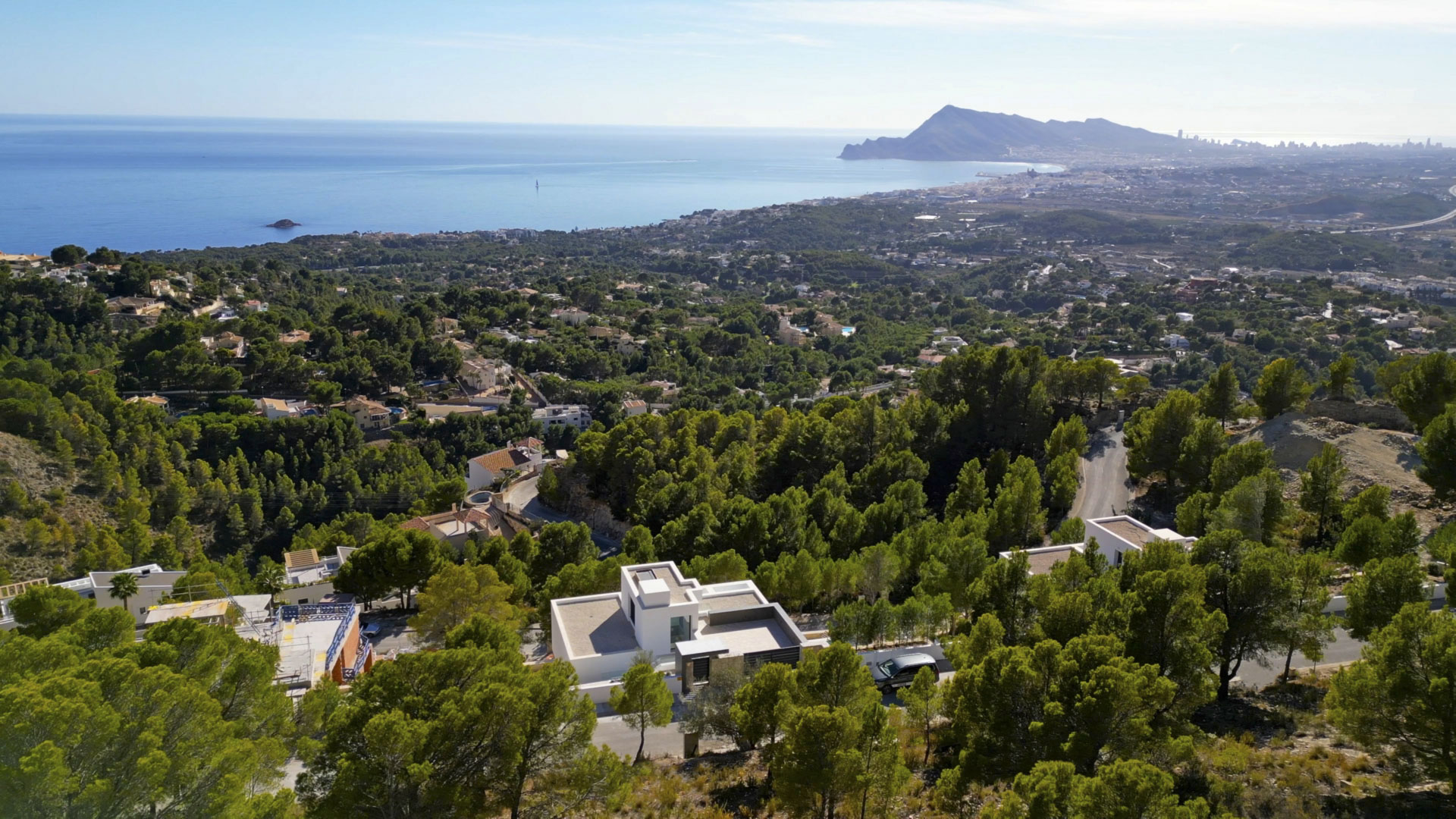Vista aérea de la villa moderna en Sierra de Altea, rodeada de pinos con vistas panorámicas al Mediterráneo y la silueta de la costa al fondo.