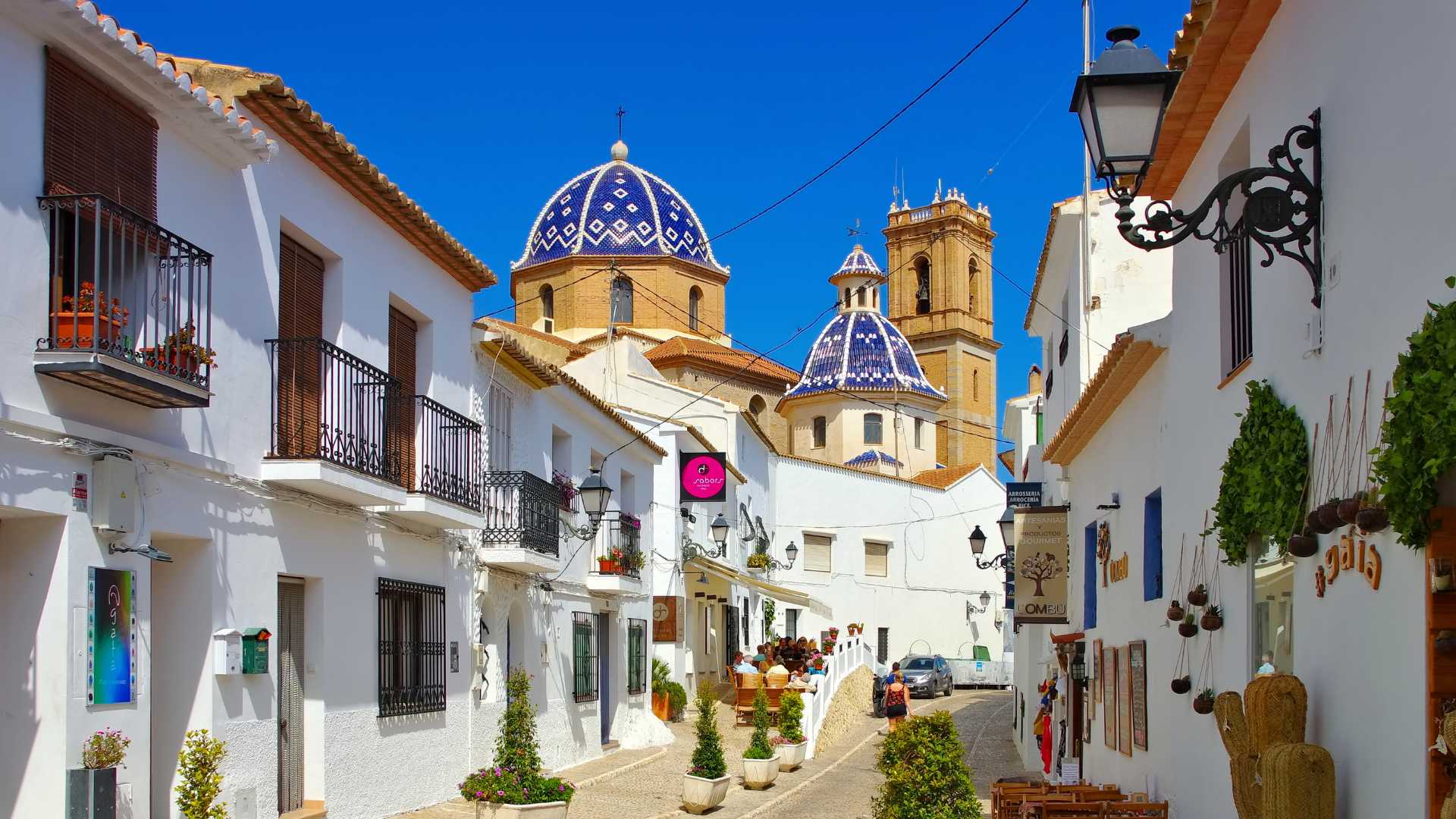 Vista del casco antiguo de Altea con fachadas blancas, balcones de forja y las icónicas cúpulas de azulejos azules de la iglesia bajo un cielo despejado.