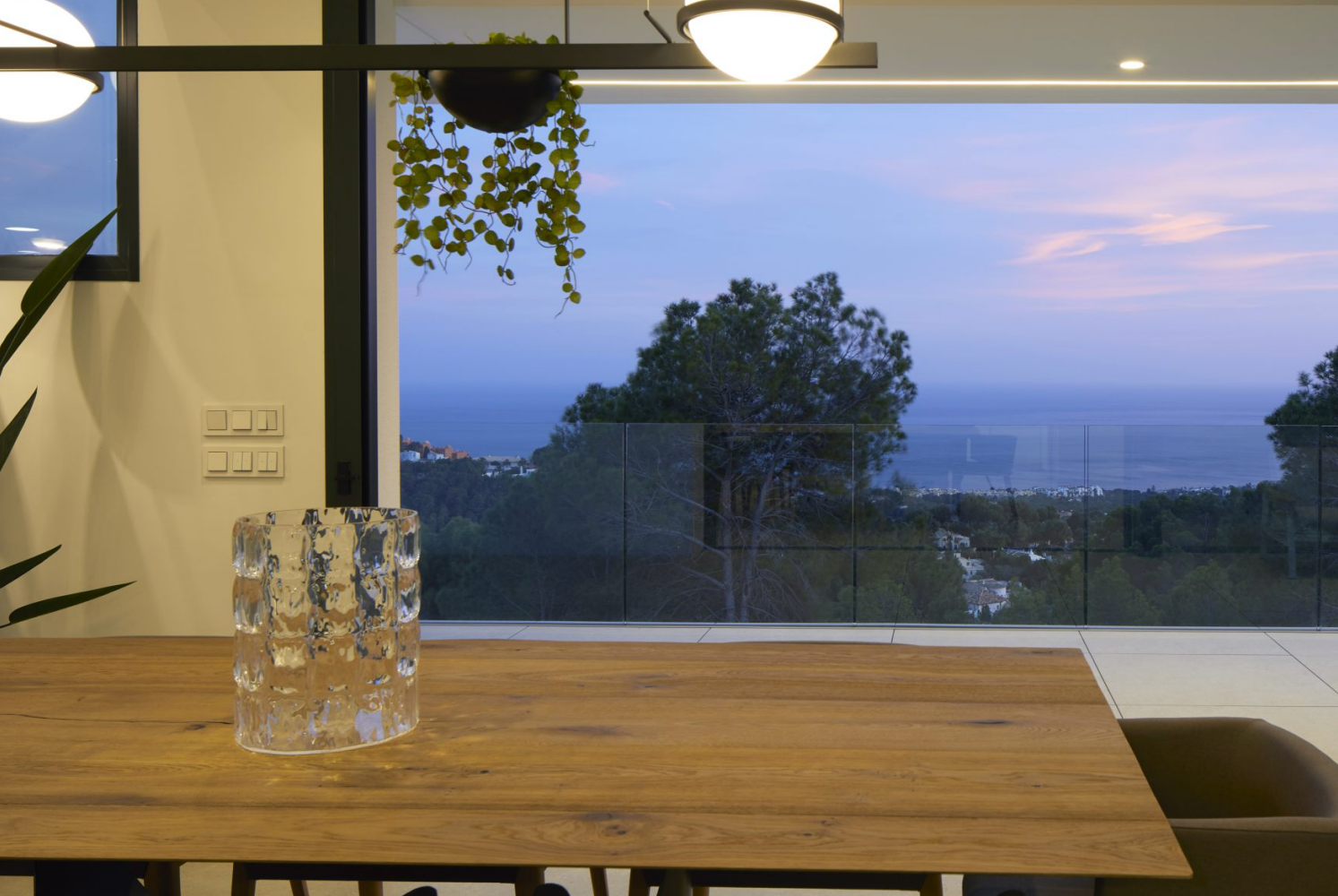 Vista desde el comedor con mesa de madera maciza hacia la terraza. El ventanal de suelo a techo ofrece vistas panorámicas al mar y la costa de Altea al atardecer.
