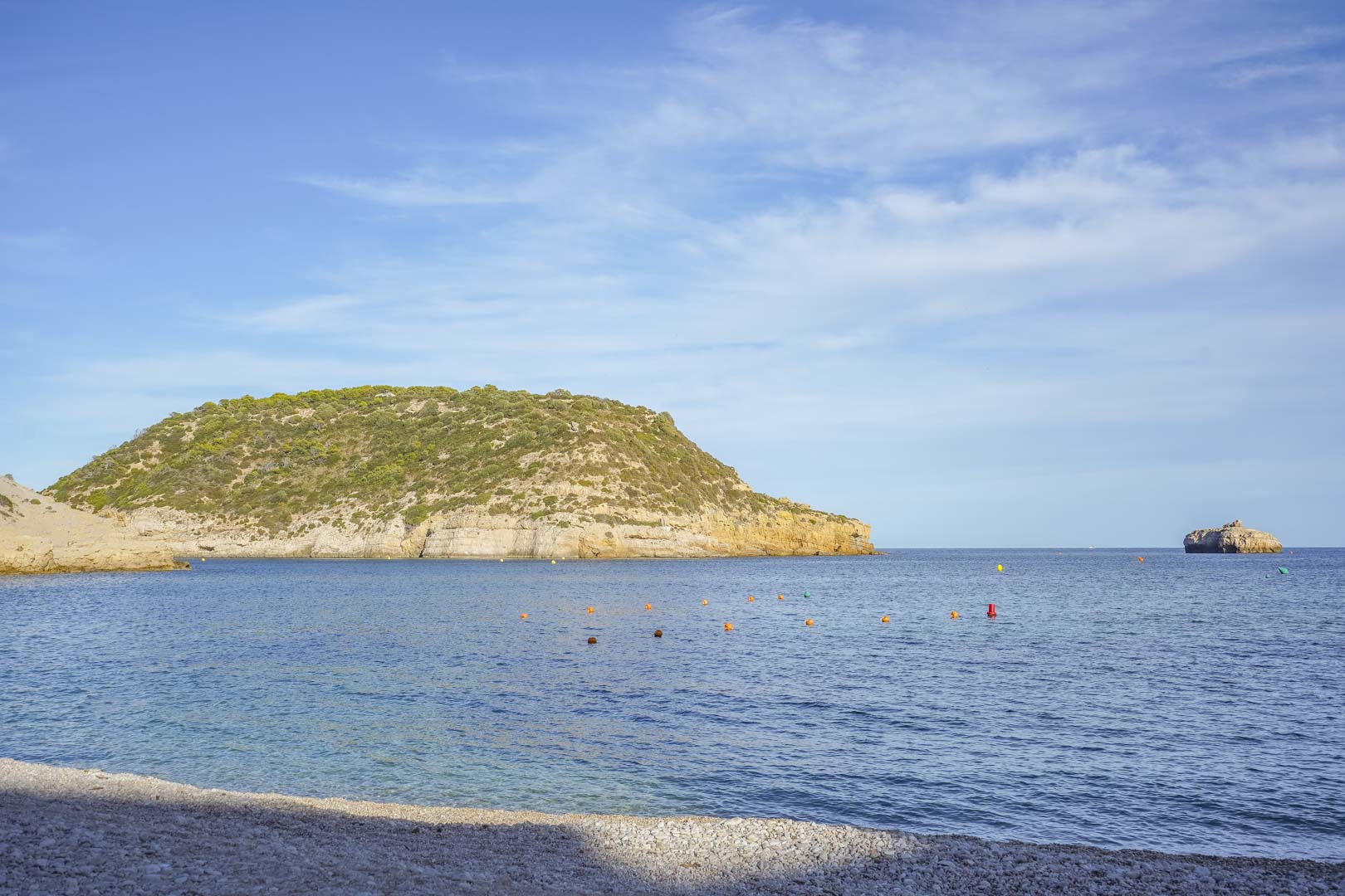 Vista panorámica de la playa de guijarros en Jávea, mostrando las aguas cristalinas del Mediterráneo y los acantilados circundantes bajo un cielo despejado.