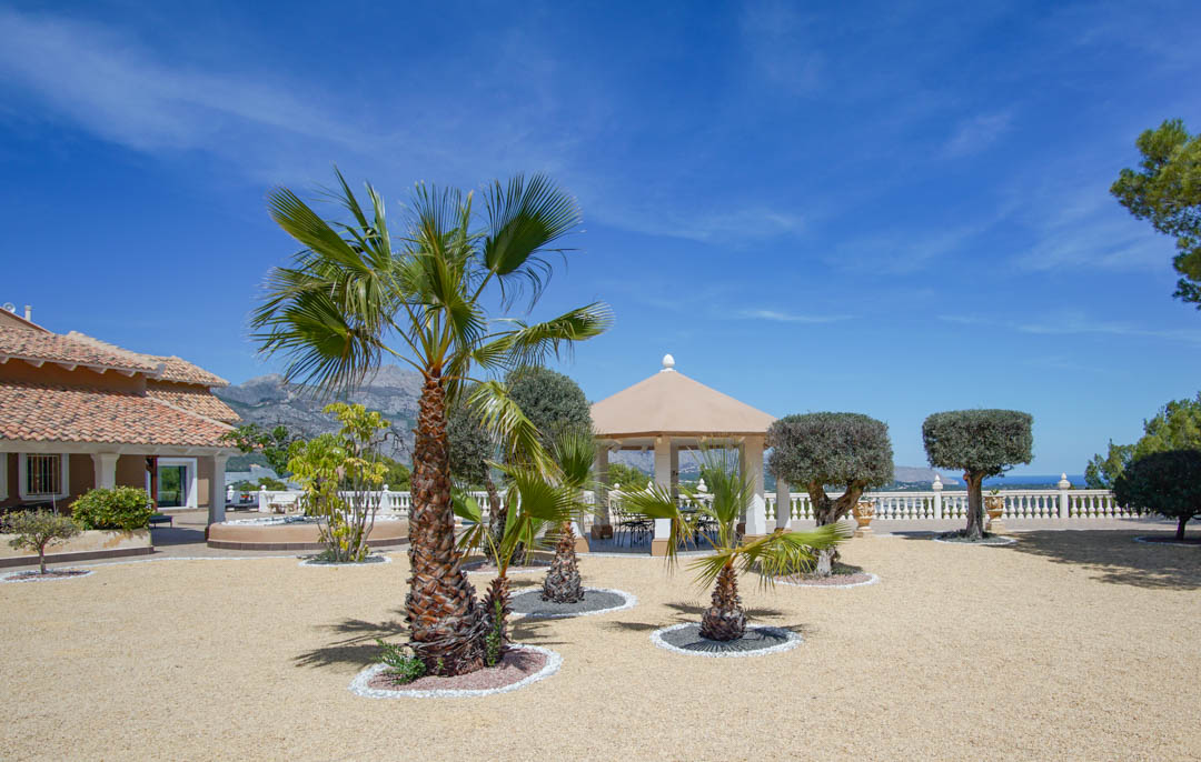 Amplio patio de grava con palmeras y olivos podados, que incluye un pabellón con columnas blancas y vistas panorámicas al mar y la montaña tras la balaustrada.