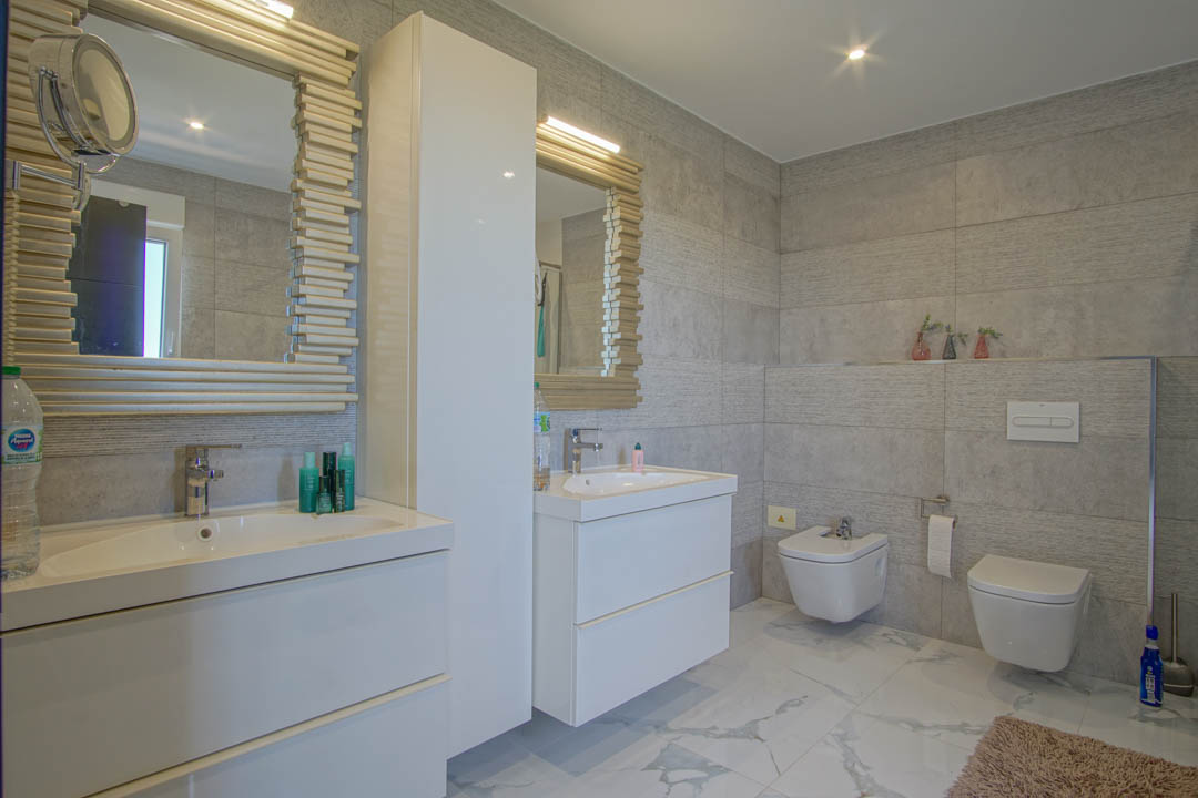Baño moderno con lavabo doble, muebles blancos suspendidos y espejos con marcos texturizados. Paredes de gres gris y suelo porcelánico efecto mármol.