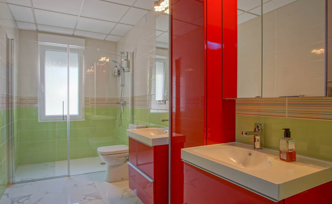 Baño moderno con mobiliario rojo brillante, lavabos dobles, ducha de obra con mampara de cristal y paredes revestidas en tonos verdes y blancos.