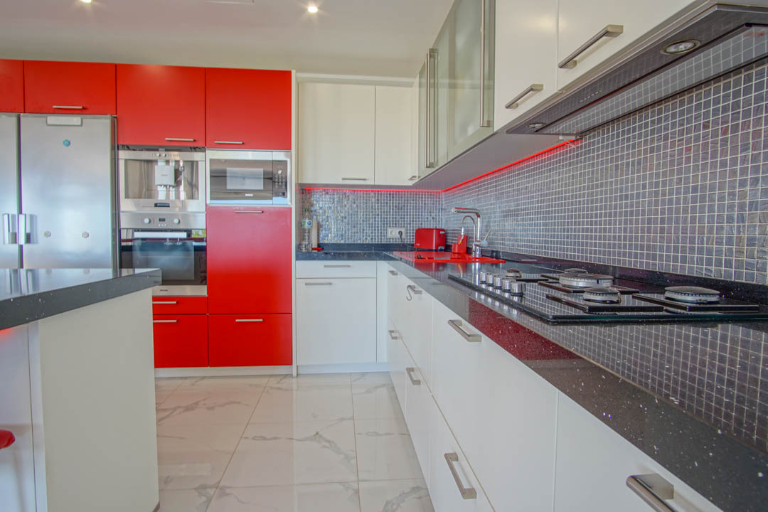 Cocina moderna con armarios en rojo y blanco brillante, encimeras de granito negro y salpicadero de mosaico gris con iluminación LED integrada.