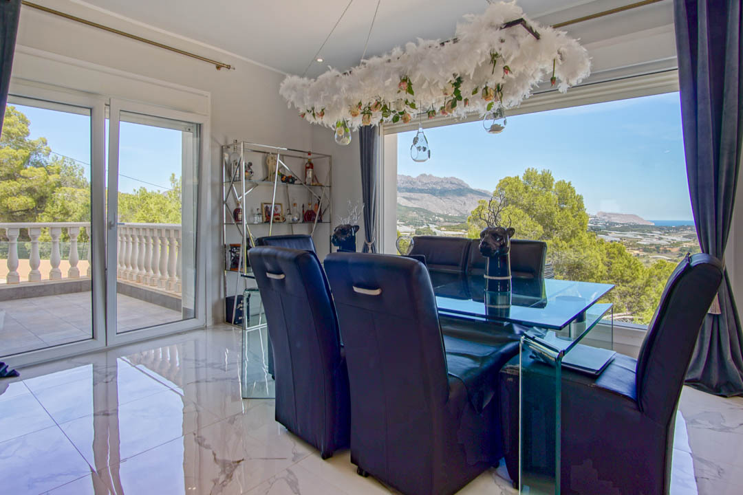 Comedor formal con suelos de mármol, mesa de cristal y sillas de cuero negro, ofreciendo vistas panorámicas a la montaña y al mar desde sus amplios ventanales.