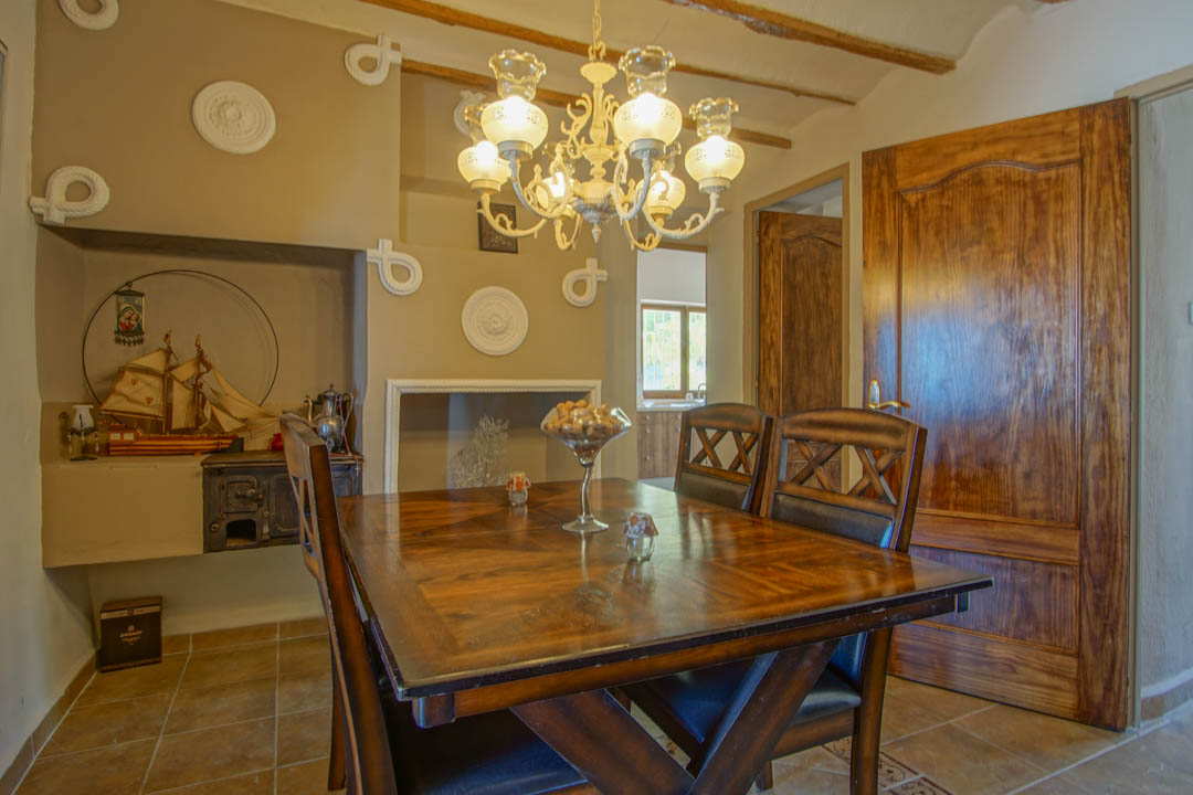 Comedor rústico con mesa de madera maciza, vigas vistas y lámpara de araña clásica. Incluye una chimenea tradicional decorada con elementos náuticos y suelos cerámicos.