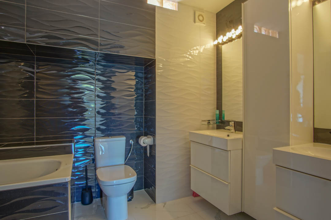 Cuarto de baño moderno con azulejos texturizados en relieve de tonos blanco y gris oscuro, mueble lavabo en blanco brillante e iluminación integrada sobre el espejo.