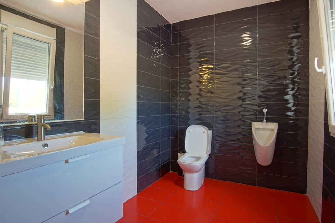 Cuarto de baño moderno con suelo de baldosas rojas, paredes de azulejos oscuros con relieve ondulado, lavabo blanco con espejo y un urinario adicional.