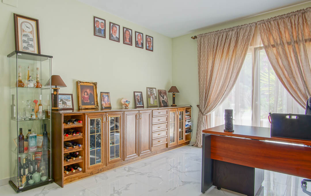 Despacho luminoso con suelo de mármol, escritorio de madera y un gran aparador con vinoteca integrada y vitrinas, iluminado por luz natural.