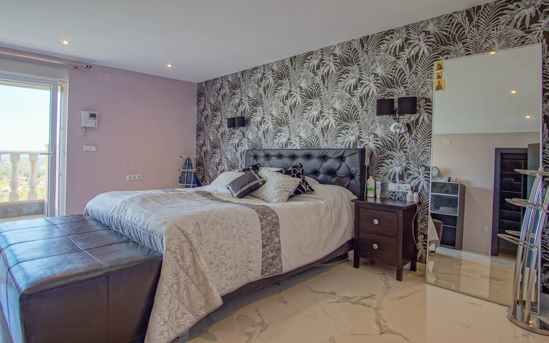 Dormitorio con suelo de mármol, pared de acento con papel tapiz tropical, cabecero de cuero capitoné y acceso directo a una terraza con balaustrada.