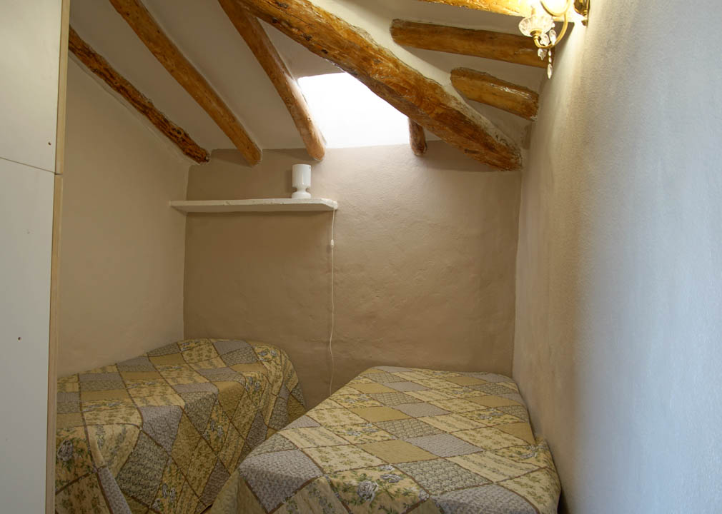 Dormitorio rústico con techos inclinados, vigas de madera a la vista y claraboya. Incluye dos camas individuales en un espacio de paredes enlucidas con acabados naturales.