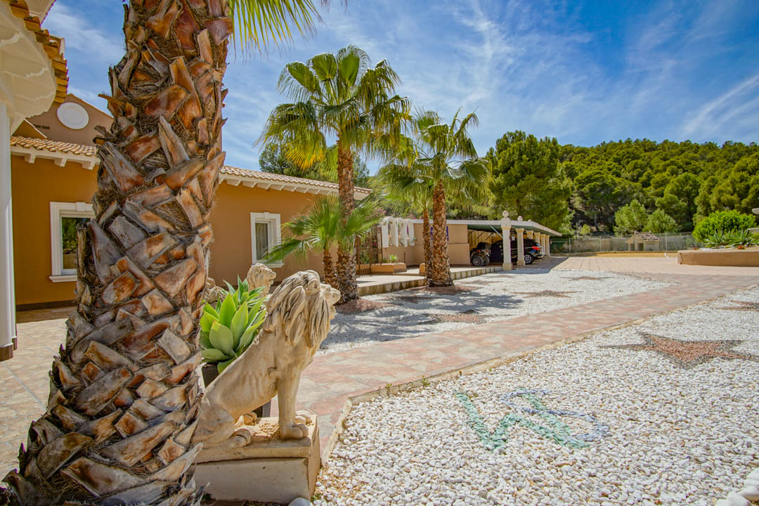 Entrada exterior con estatuas de leones de piedra, palmeras y un camino de grava decorado con mosaicos que conduce a una cochera cubierta para varios vehículos.