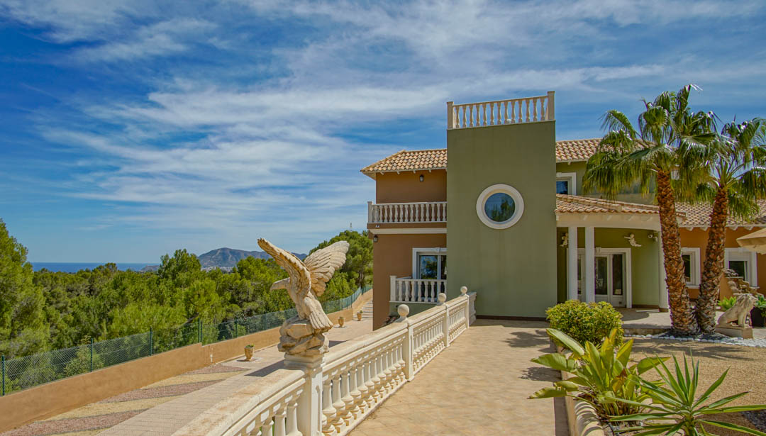 Fachada exterior con balaustradas clásicas, escultura ornamental de águila y entrada con palmeras que ofrece vistas panorámicas al bosque de pinos y al mar Mediterráneo.