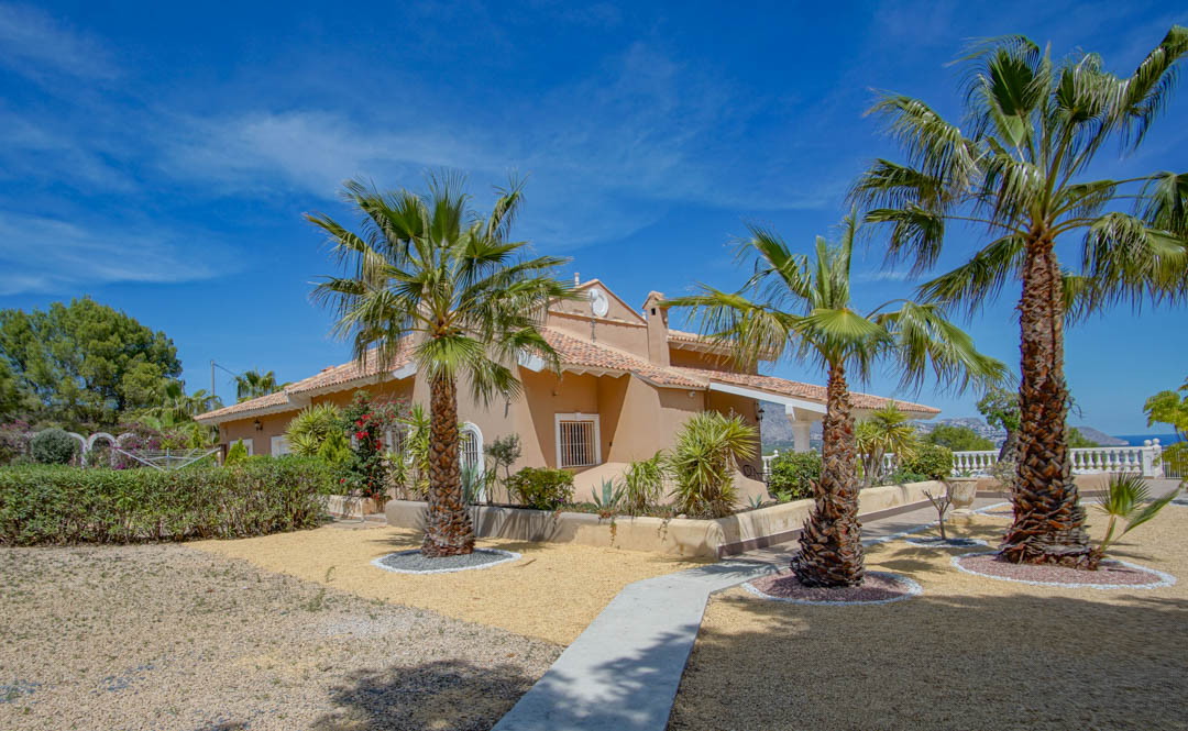 Fachada mediterránea de tonos tierra con tejado de teja, rodeada de jardines de grava con palmeras y senderos pavimentados bajo un cielo despejado.