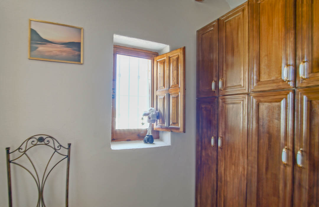 Habitación rústica con armario empotrado de madera maciza, ventana tradicional con contraventana de madera y silla de forja decorativa.