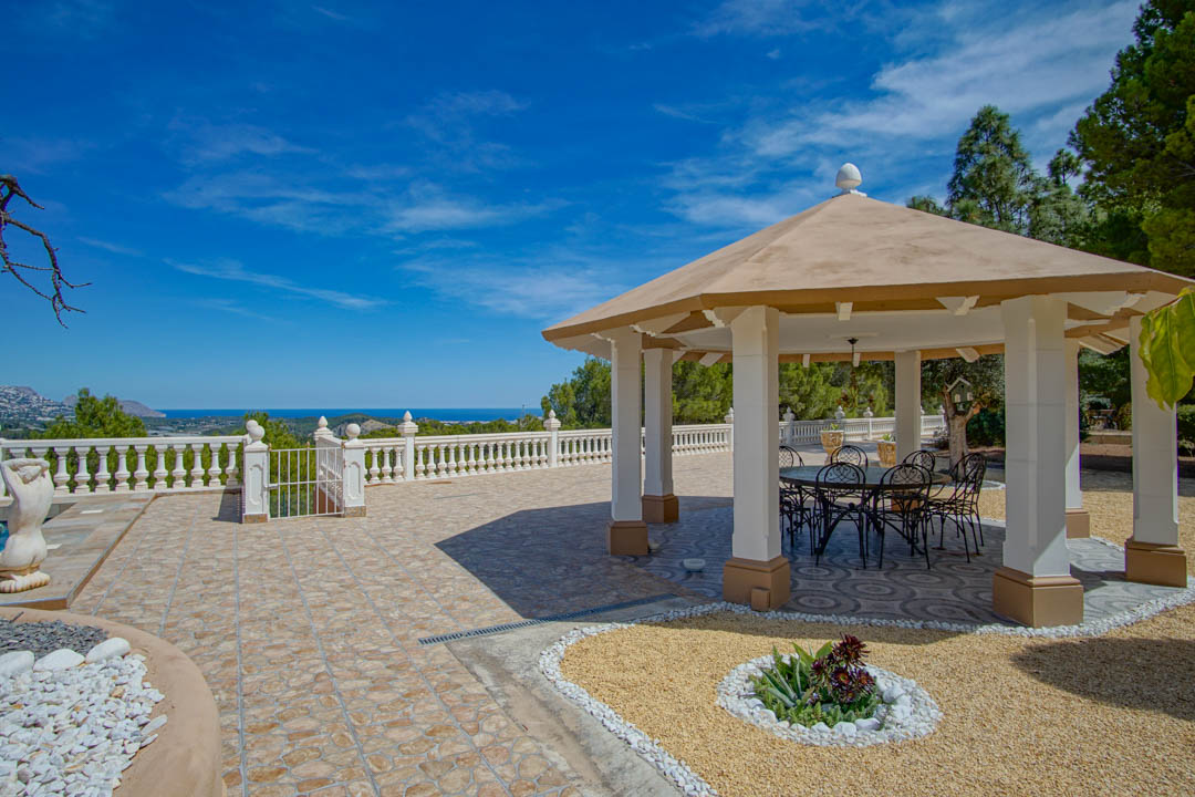 Pabellón exterior con columnas blancas y mobiliario de hierro forjado, situado sobre una terraza de baldosas con vistas panorámicas al mar Mediterráneo.