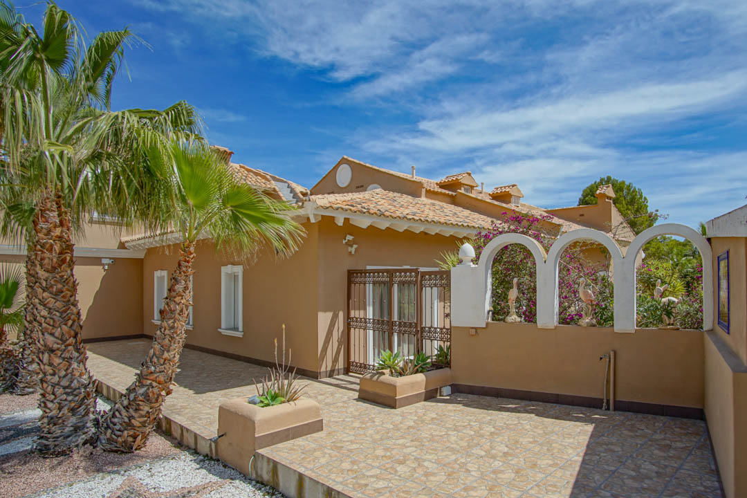 Patio exterior con suelos cerámicos y muros ocre, destacando arcos decorativos blancos con figuras ornamentales, palmeras y una puerta de forja tradicional.