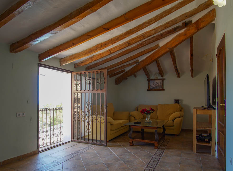 Sala de estar rústica con vigas de madera a la vista, suelos de gres y acceso a un balcón a través de una puerta con rejería ornamental.