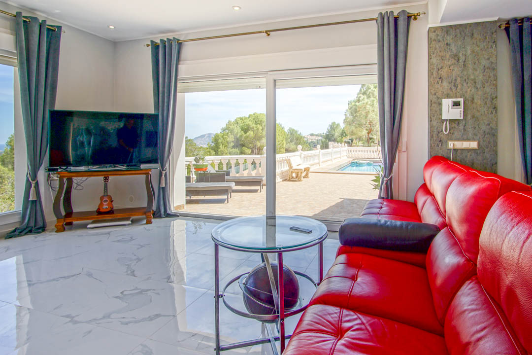 Salón con suelos de mármol blanco, sofá de cuero rojo y grandes ventanales que ofrecen acceso directo a la terraza con piscina y vistas despejadas.