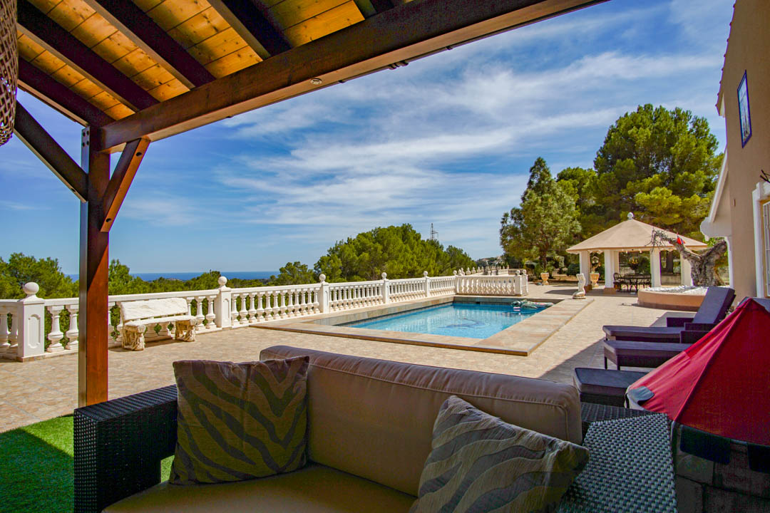 Terraza con piscina, balaustrada blanca y vistas panorámicas al mar. Cuenta con una zona de estar bajo pérgola de madera y un pabellón entre pinos mediterráneos.
