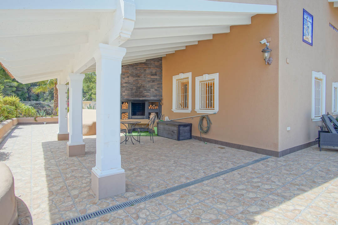Terraza cubierta con vigas de madera blanca, columnas clásicas y zona de barbacoa integrada con chimenea de piedra y almacenamiento de leña.