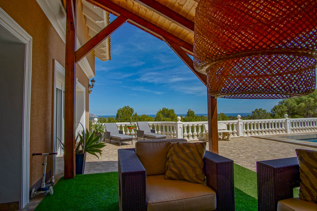 Terraza cubierta con vigas de madera y zona de estar, con vistas panorámicas al mar Mediterráneo tras una balaustrada clásica blanca.