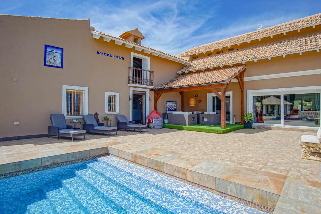 Terraza principal con piscina de mosaico azul, tumbonas y un porche cubierto con vigas de madera que alberga una zona de estar sobre césped artificial.