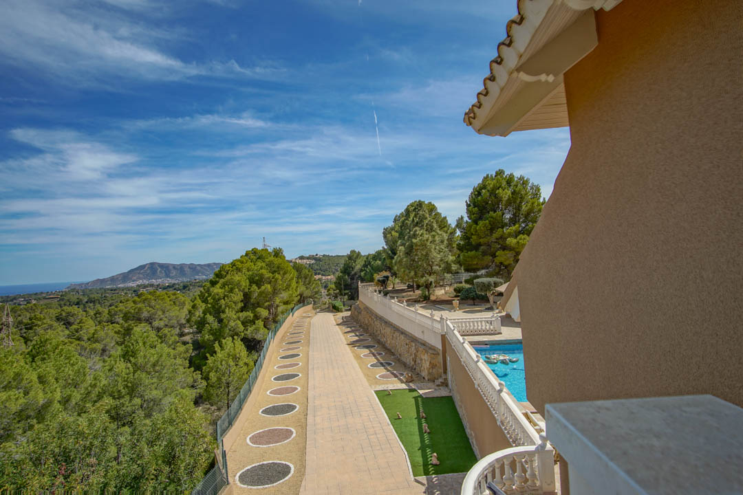 Vista elevada que muestra el camino pavimentado, la zona de piscina con balaustrada blanca y el entorno natural con vistas panorámicas al mar y la montaña.