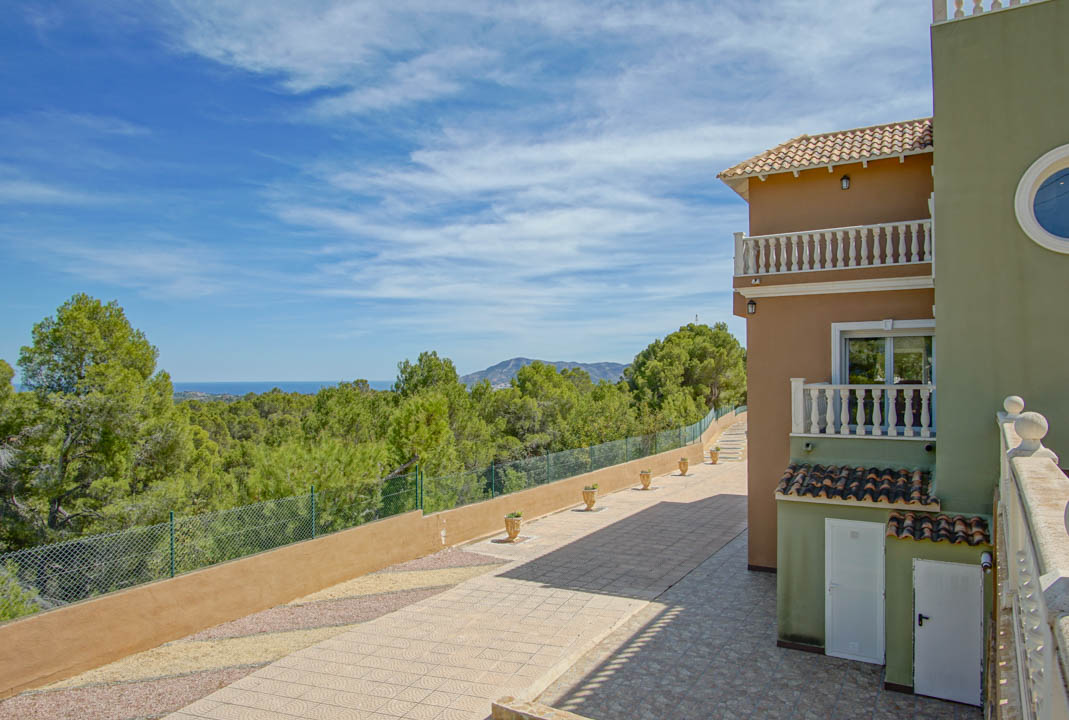 Vista exterior de la fachada con balcones de balaustrada, camino pavimentado y vistas panorámicas despejadas hacia el bosque de pinos y el mar Mediterráneo.