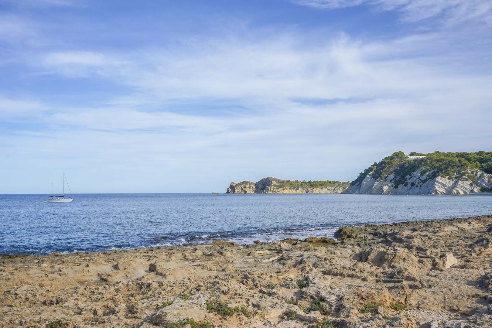 Vista de la costa rocosa de Jávea con el mar Mediterráneo, un velero navegando y acantilados con vegetación bajo un cielo despejado.