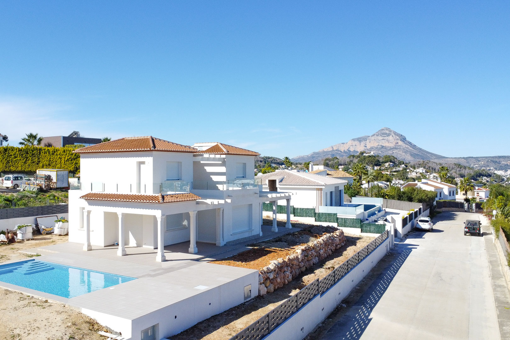 Vista exterior de la villa blanca con tejado de teja cerámica, piscina integrada y muros de piedra natural, destacando la montaña del Montgó al fondo.