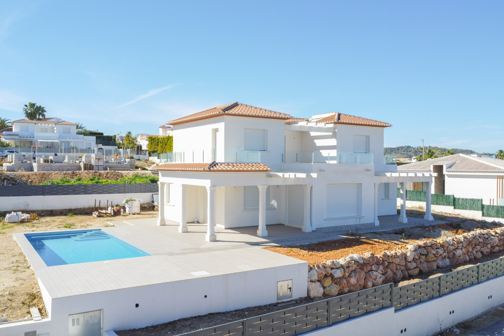 Vista exterior de la villa blanca en fase final de obra, con piscina rectangular, porches con columnas y terrazas con barandillas de cristal bajo el cielo despejado.