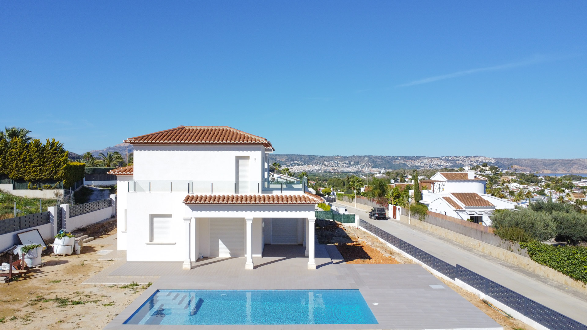 Vista exterior de la villa en fase final de obra, destacando la piscina rectangular, la fachada blanca de dos niveles y las terrazas con barandillas de cristal.