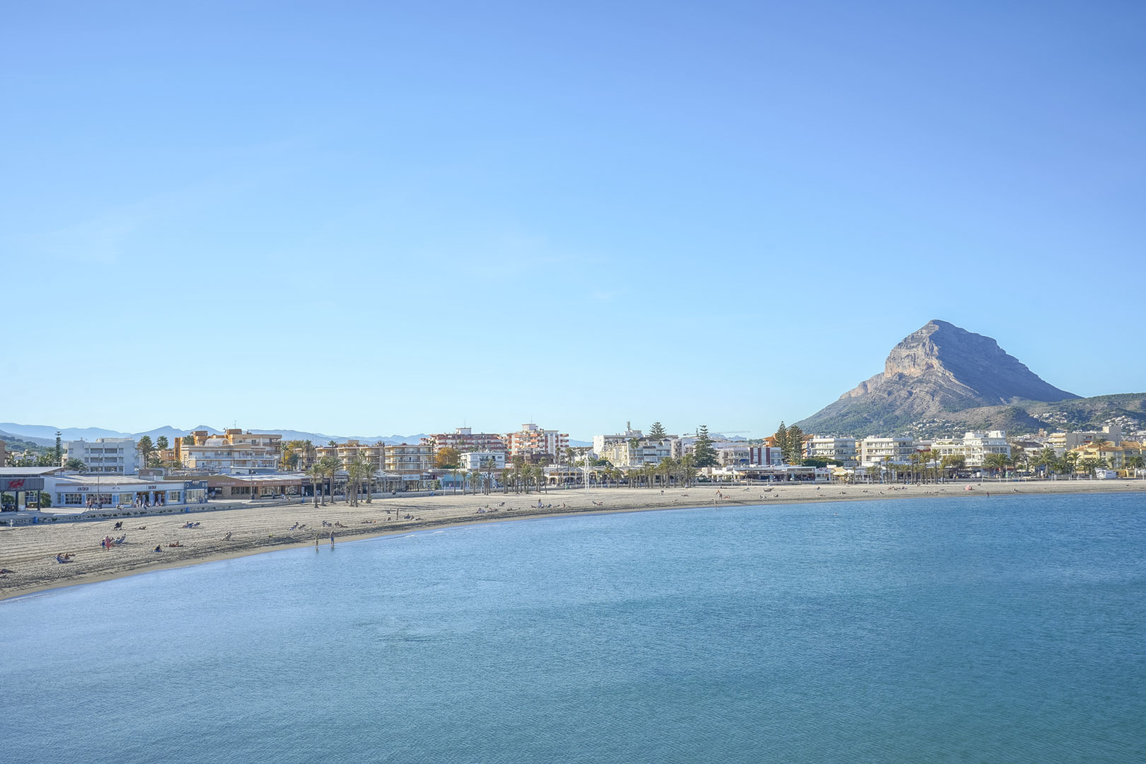 Vista panorámica de la playa del Arenal en Jávea, destacando su costa de arena fina y el emblemático monte Montgó al fondo bajo un cielo despejado.