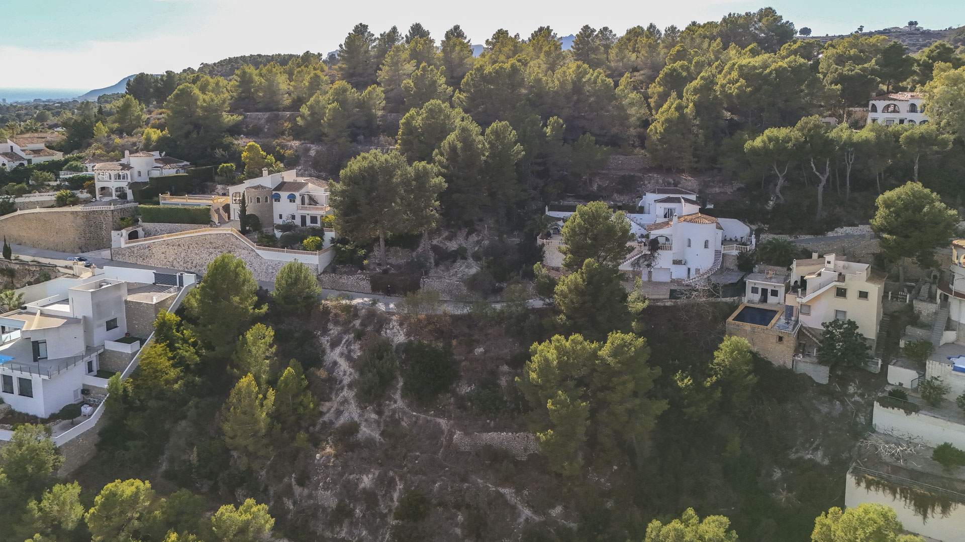 Vista aérea de parcelas residenciales en Benissa, rodeadas de densa vegetación de pinos y muros de piedra natural, con el mar Mediterráneo al fondo.