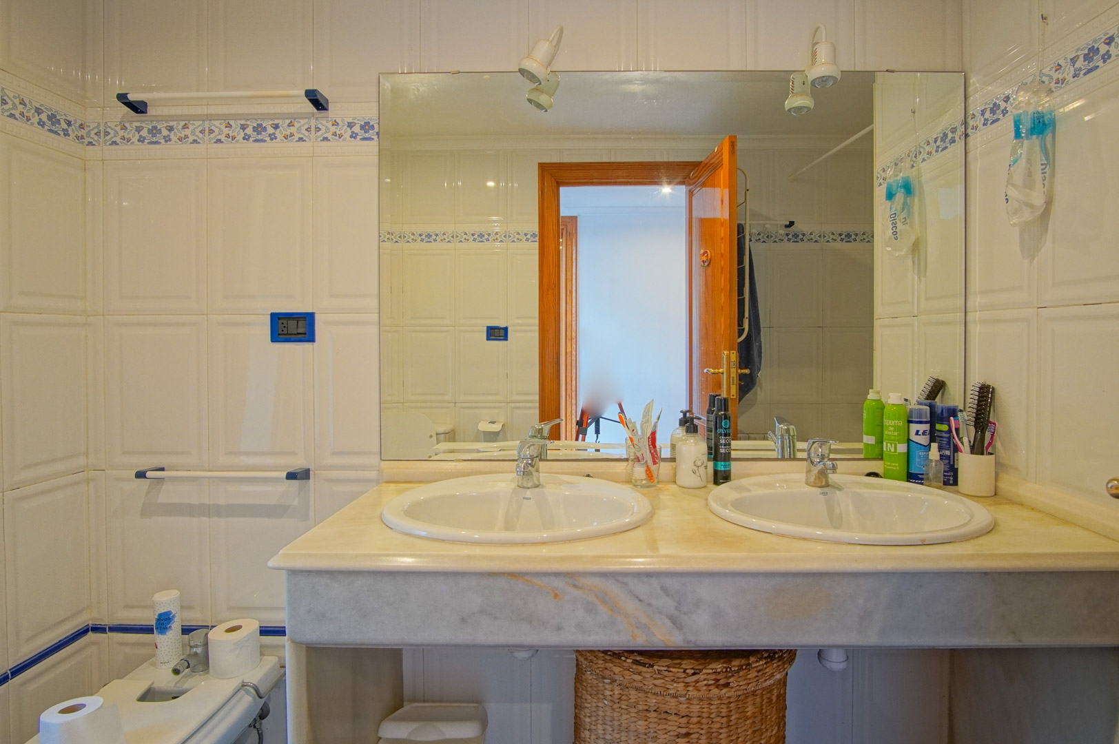 Baño con encimera de mármol y lavabo de doble seno, paredes alicatadas en blanco con cenefa floral azul y un gran espejo con iluminación integrada.