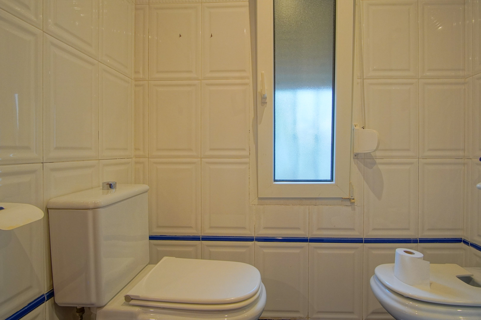 Baño con paredes alicatadas en blanco con relieve y cenefa azul. Incluye inodoro, bidé y una ventana de cristal translúcido que aporta luz natural.