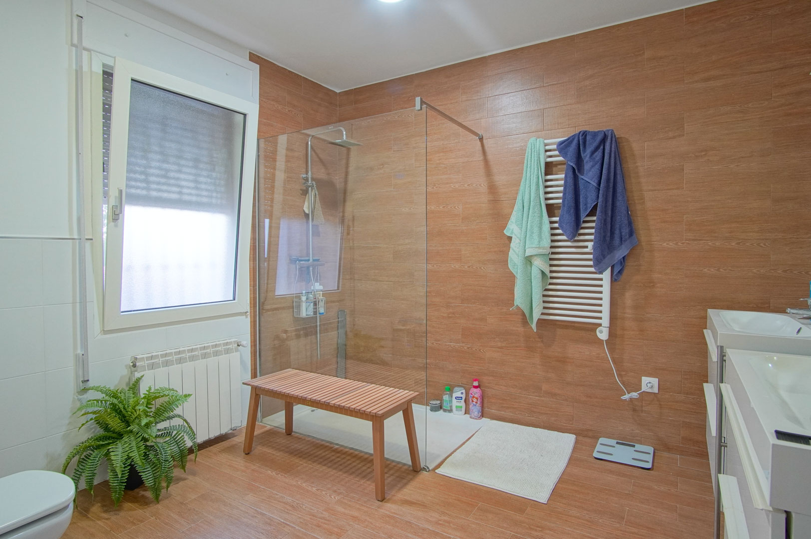 Baño moderno con azulejos de efecto madera, ducha a ras de suelo con mampara de cristal, doble lavabo blanco y radiador toallero eléctrico.