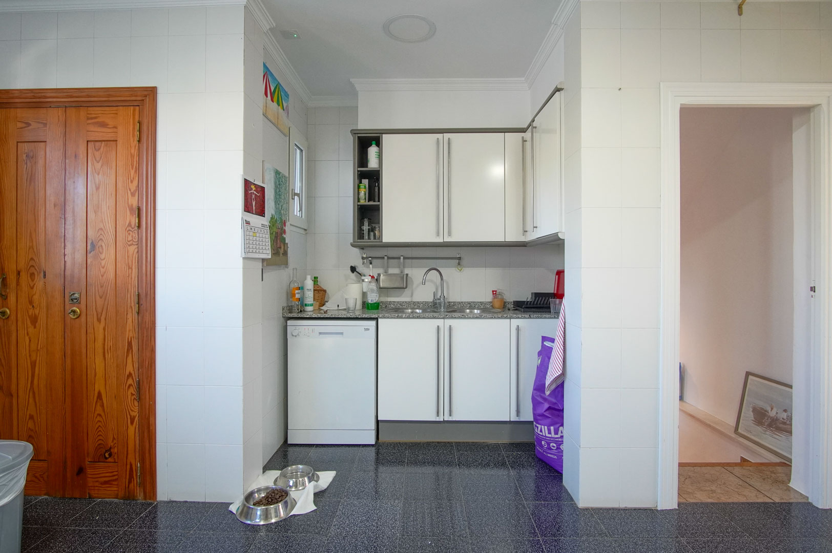 Cocina funcional del apartamento de invitados con armarios blancos, encimera de granito y suelos de terrazo oscuro, situada junto al acceso a las plantas superiores.