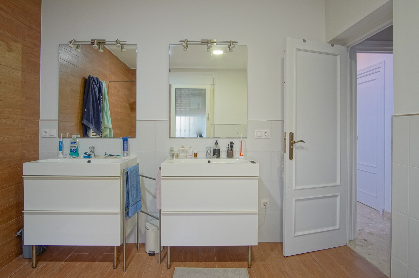 Cuarto de baño con lavabo doble independiente, espejos con iluminación superior y revestimiento de pared que combina azulejos blancos con gres de efecto madera.