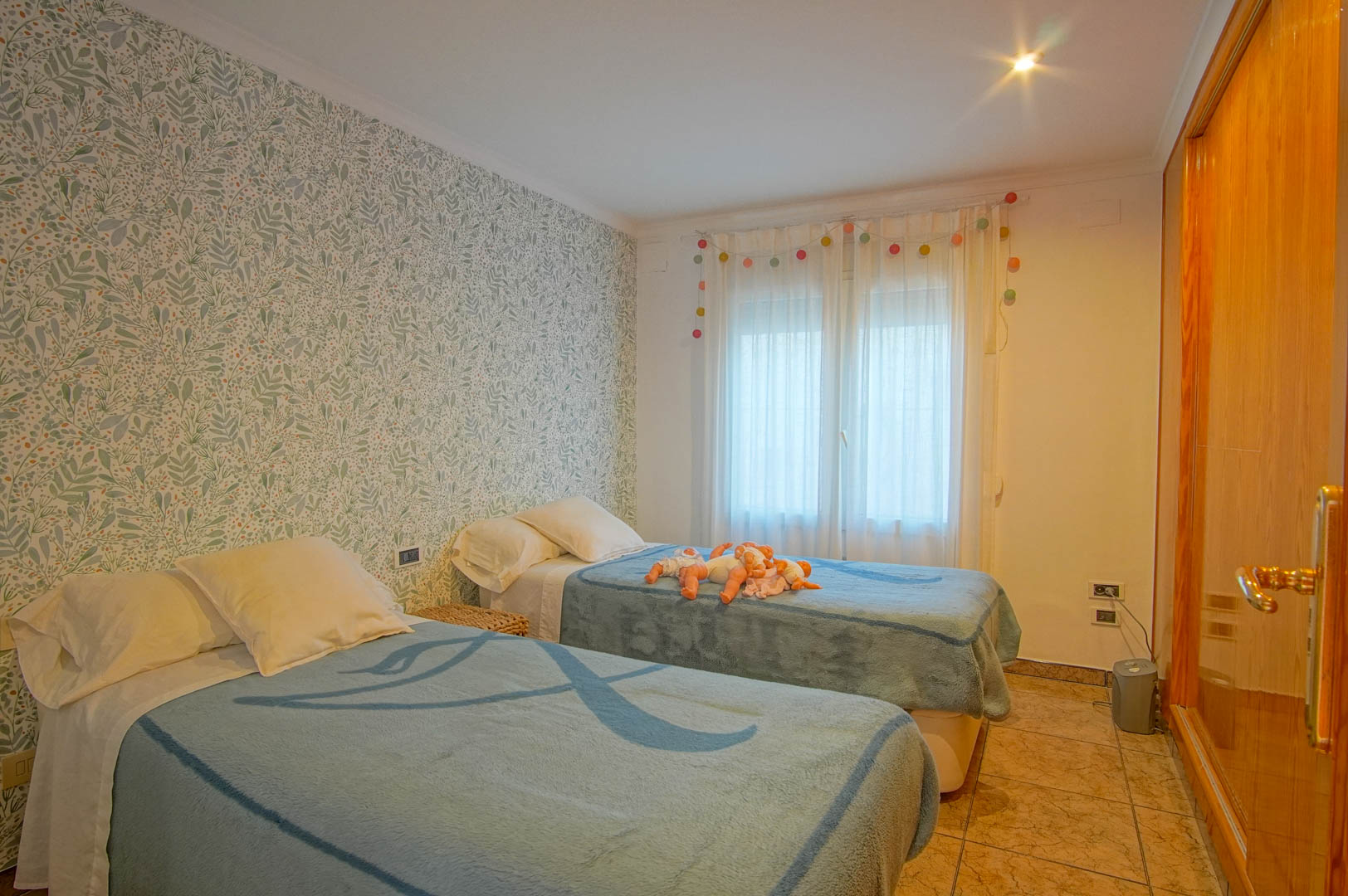Dormitorio con dos camas individuales, pared con papel pintado de motivos botánicos, armario empotrado de madera y suelos de gres.