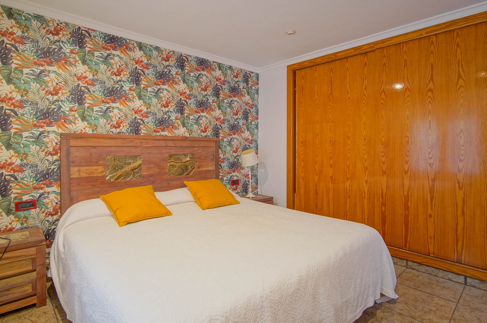 Dormitorio con papel pintado de motivos tropicales, cabecero de madera con incrustaciones de piedra y un amplio armario empotrado de madera natural.
