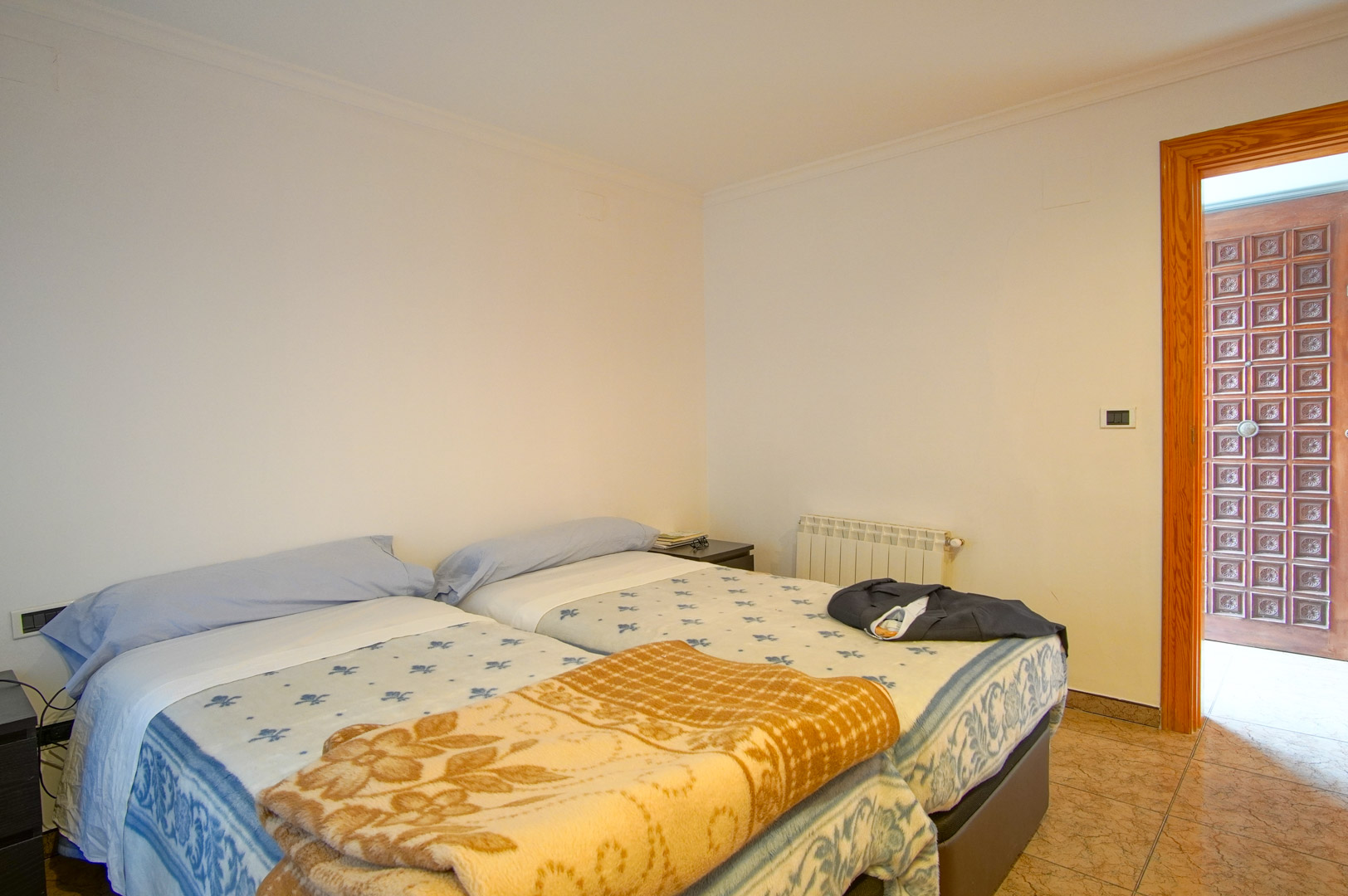 Dormitorio funcional con dos camas individuales, paredes blancas y suelos cerámicos. Incluye calefacción por radiador y una puerta de madera con paneles tallados.