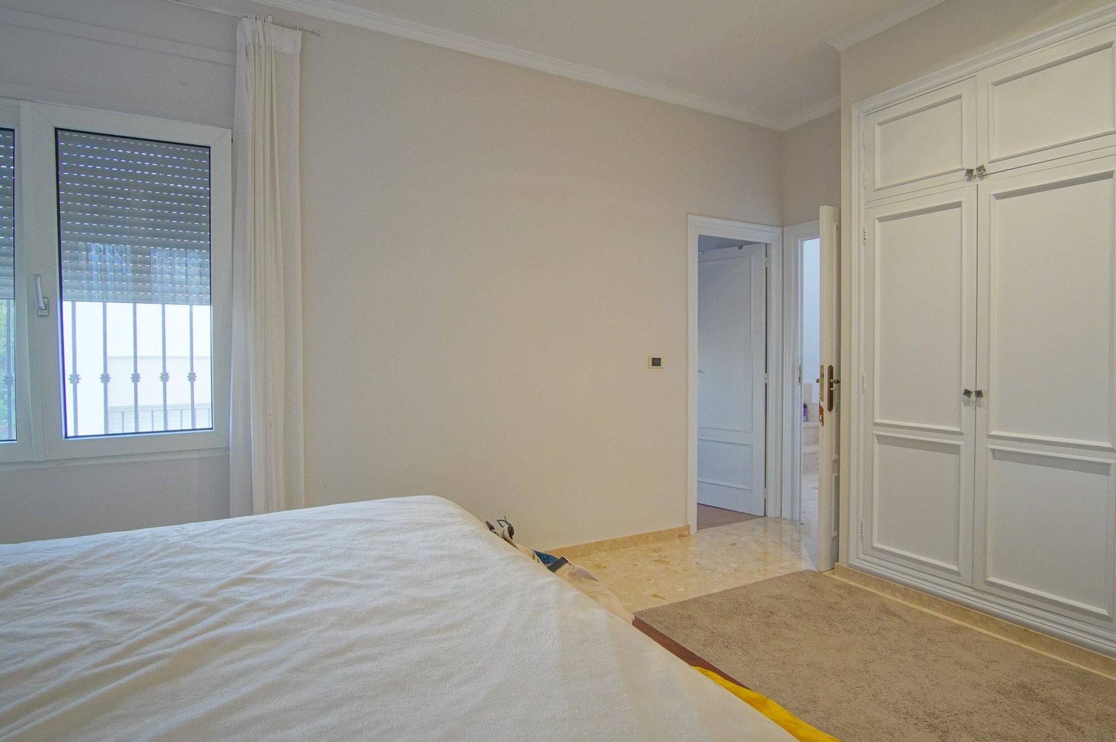 Dormitorio luminoso con armarios empotrados blancos, suelos de mármol y ventana con persiana. El diseño neutro ofrece un ambiente tranquilo y funcional.
