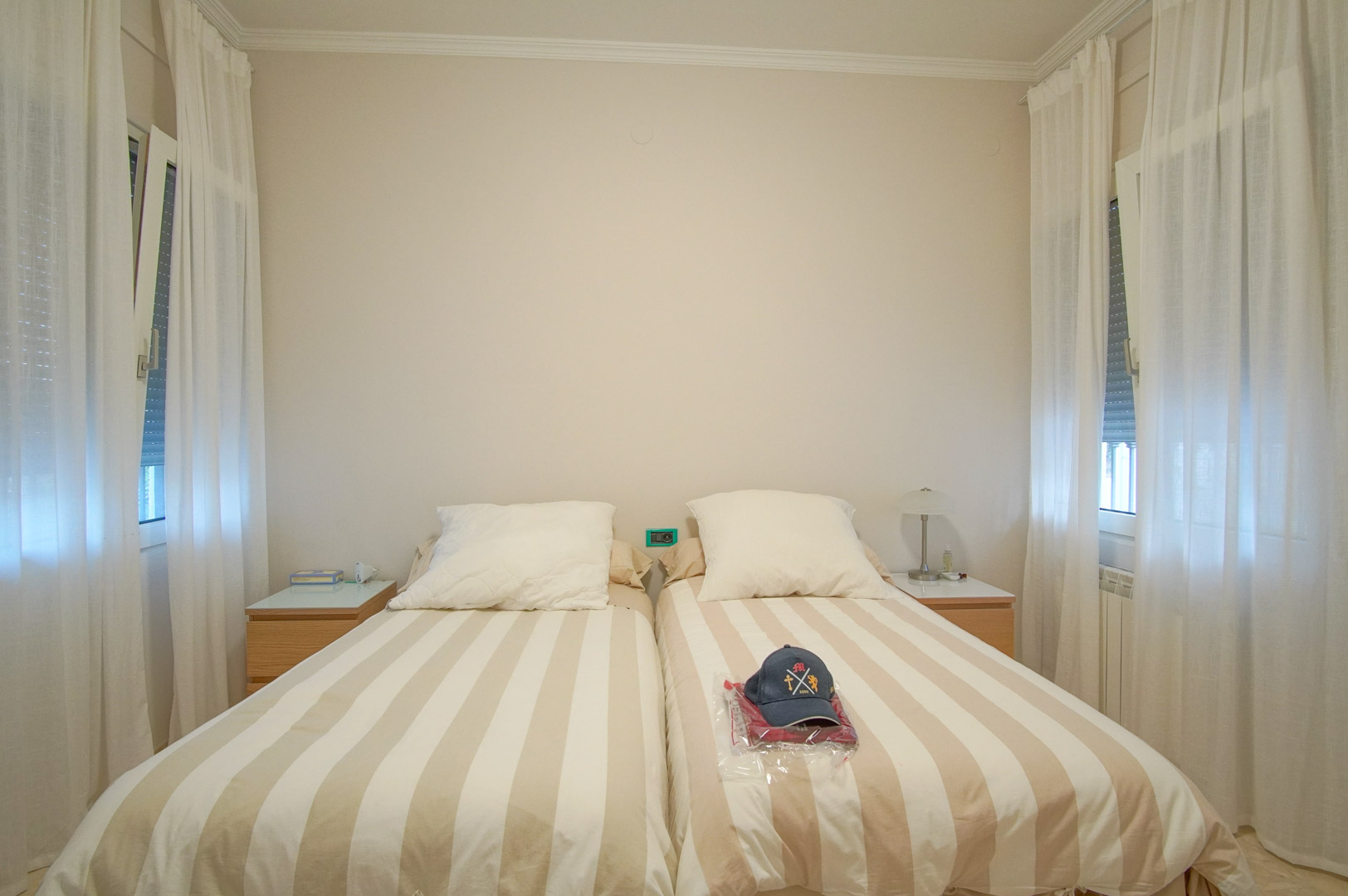 Dormitorio luminoso con dos camas individuales, mesitas de noche de madera y ventanas laterales con cortinas blancas que aportan luz natural y ventilación.