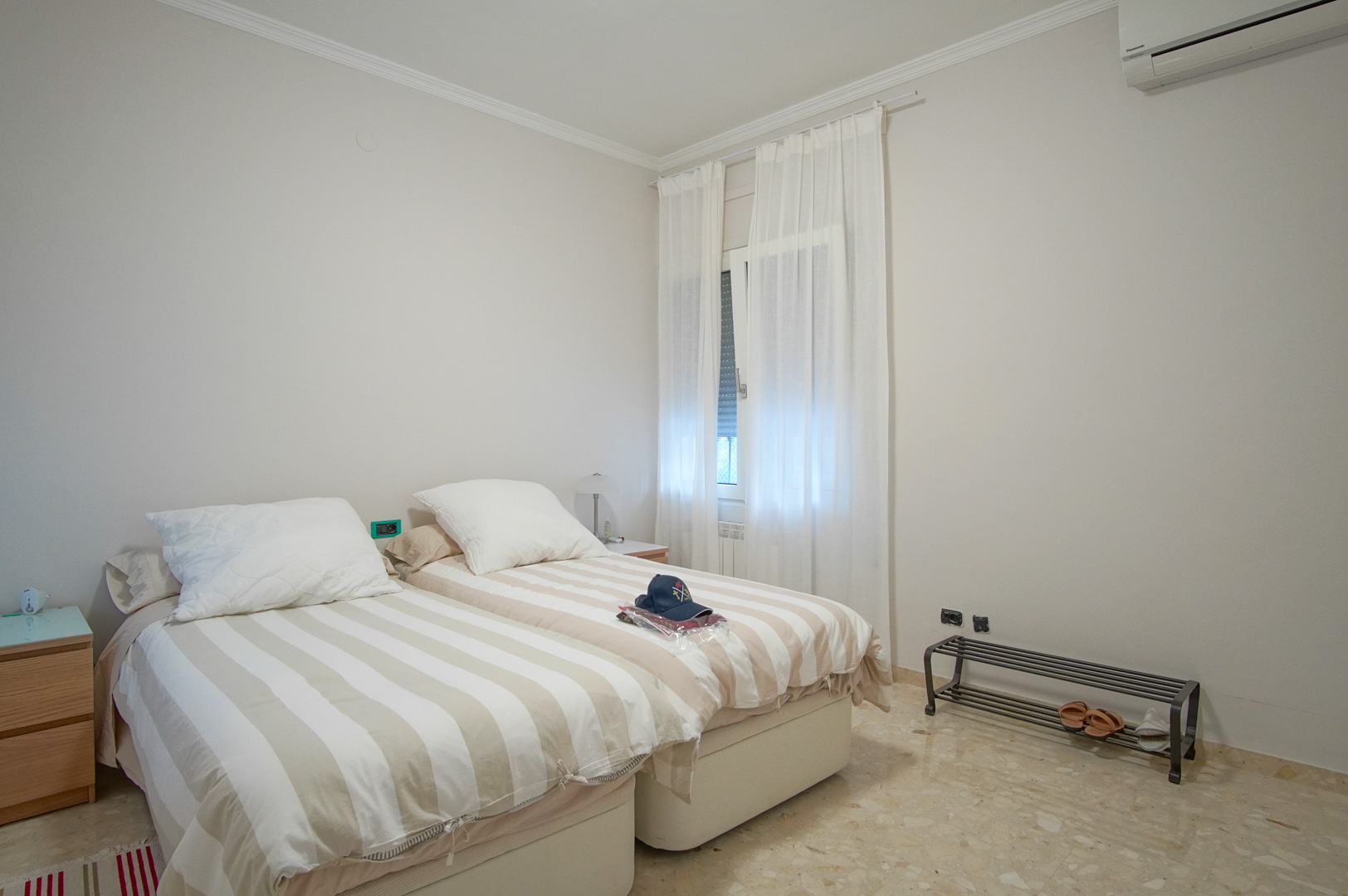 Dormitorio luminoso con dos camas individuales, suelos de mármol, aire acondicionado y una ventana con cortinas blancas que aporta luz natural.