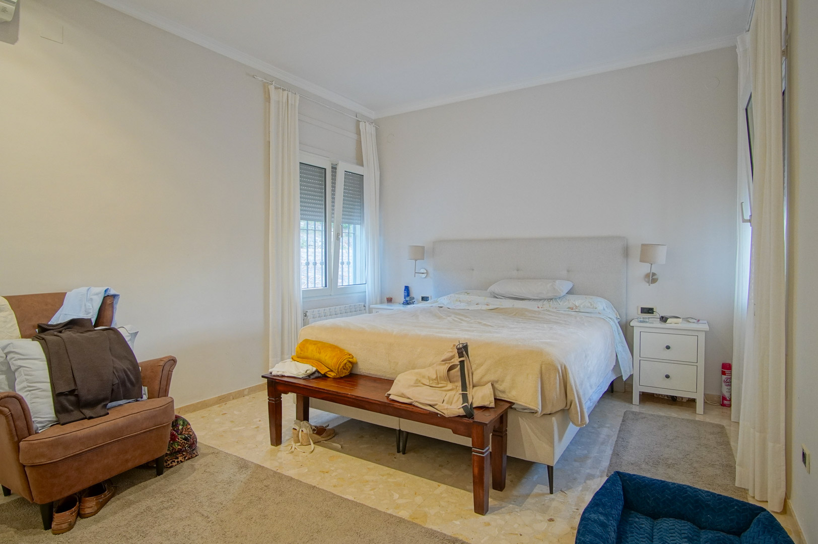 Dormitorio luminoso con suelos de mármol, cama doble con cabecero tapizado y banco de madera. Incluye ventanales con persianas y acceso a luz natural.